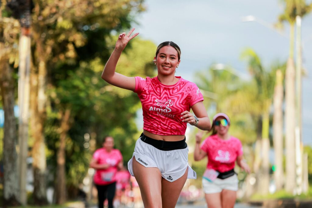 Energetic woman running outdoors in pink event shirt, smiling and flashing a peace sign.